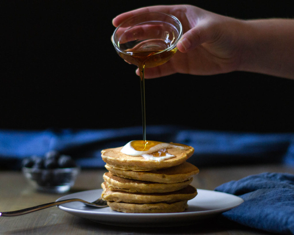 maple syrup being poured onto a stack of sweet potato pancakes with blueberries in the background