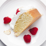 A slice of almond pound cake on a white plate, topped with whipped cream, sliced toasted almonds, and fresh raspberries. Text reads "easy almond poundcake" and "ficklebeabkehouse.com"