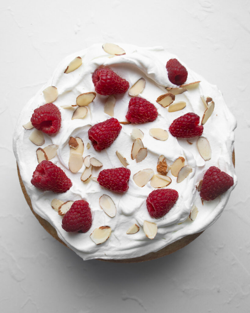 Almond pound cake shown from above and topped with whipped cream, sliced almonds, and fresh raspberries.