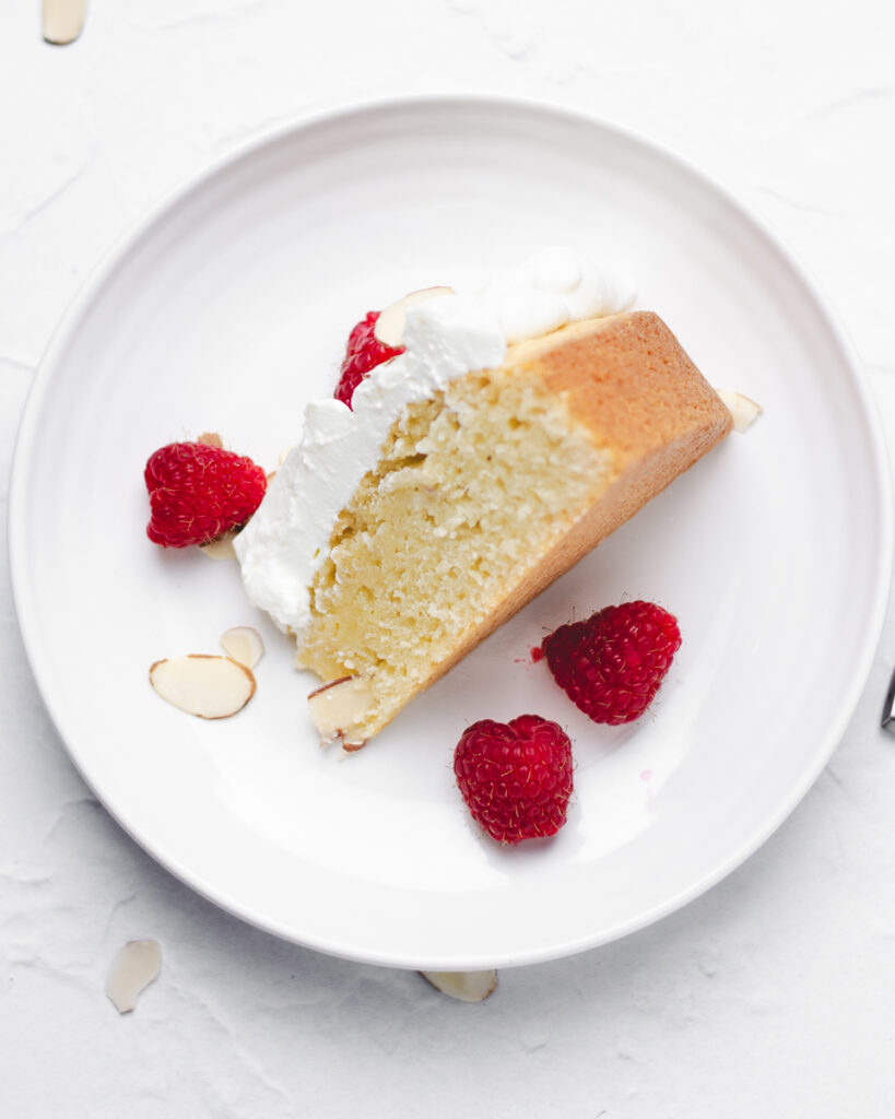 Slice of almond pound cake shown from above with fresh whipped cream, sliced almonds, and fresh raspberries.